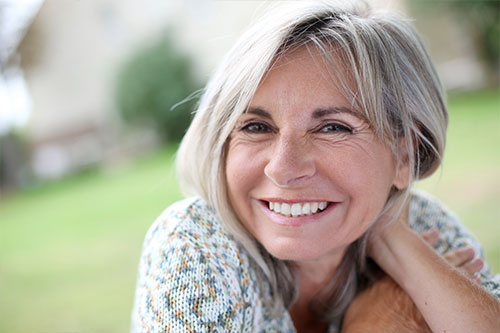 Older woman with grey hair smiling at the camera with a blurred out park scene in the background