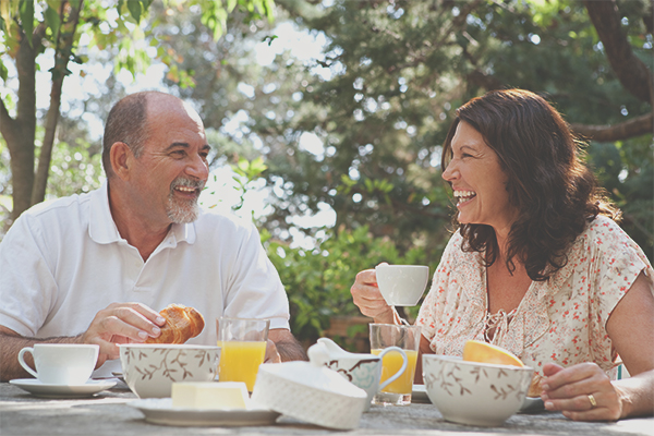 Older couple smiling while enjoying coffee, juice, and croissants at a table outside with trees in the background
